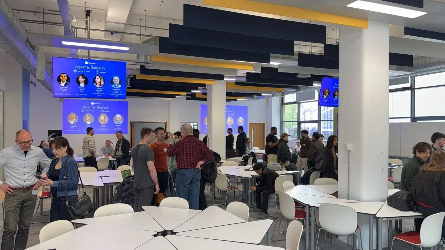 Guests standing and talking among white tables in a bright open room at Agentic Society, LSE, with blue screens on the walls showing the event name.