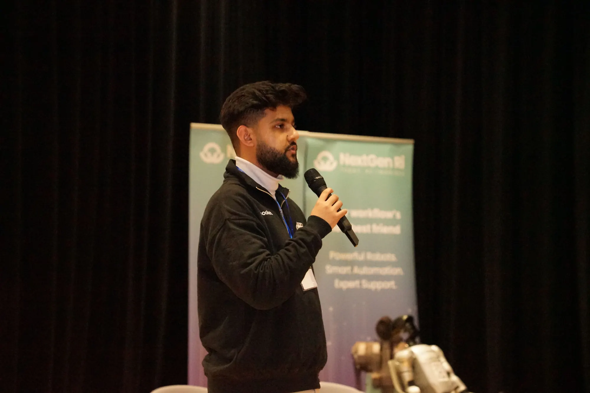 Abdullah with a microphone on stage at King's College London, in front of a NextGenRI banner.