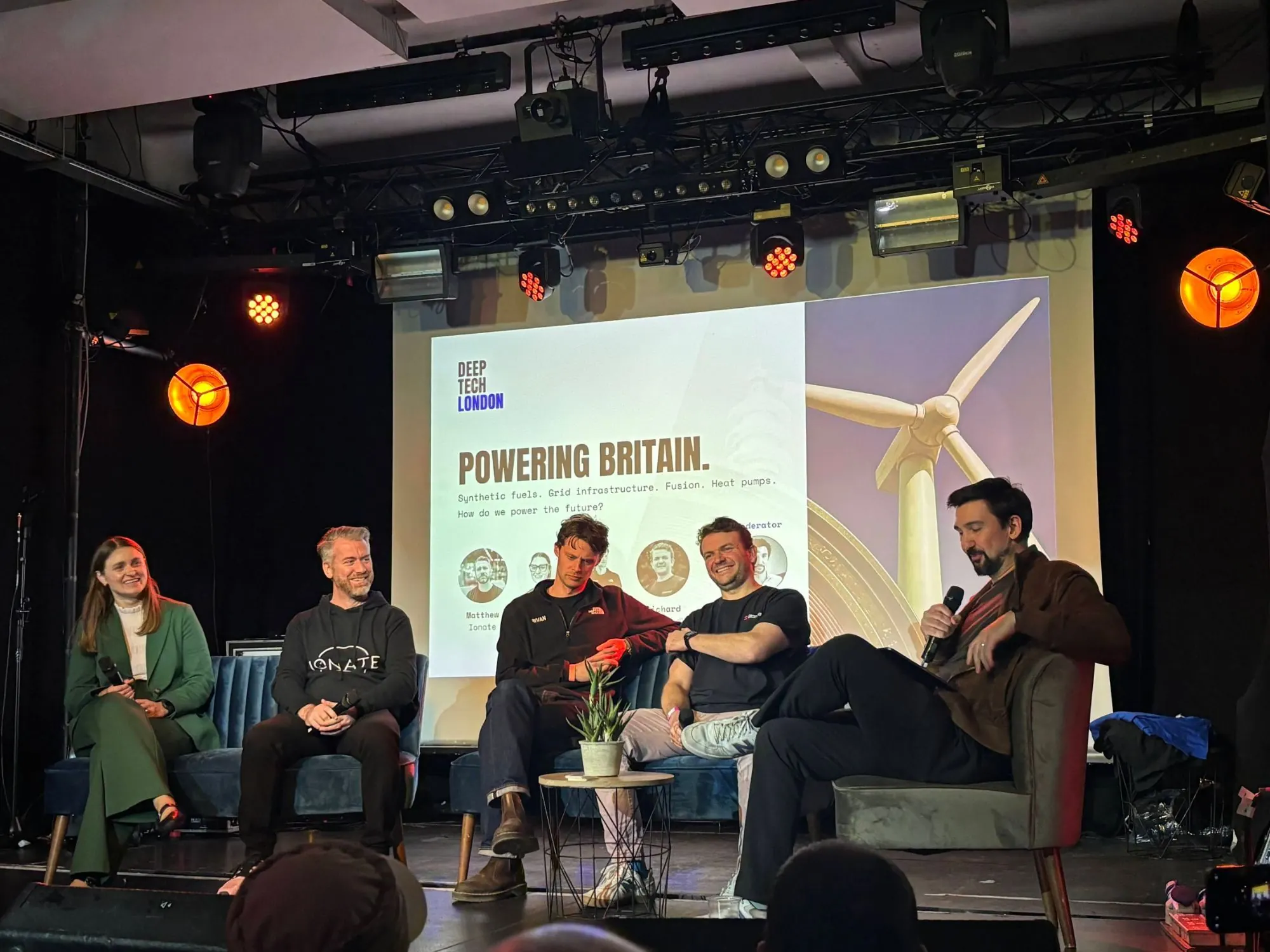 Five people seated on a stage in front of a Deep Tech London slide titled "Powering Britain" with a wind turbine on it, one of them speaking into a microphone.