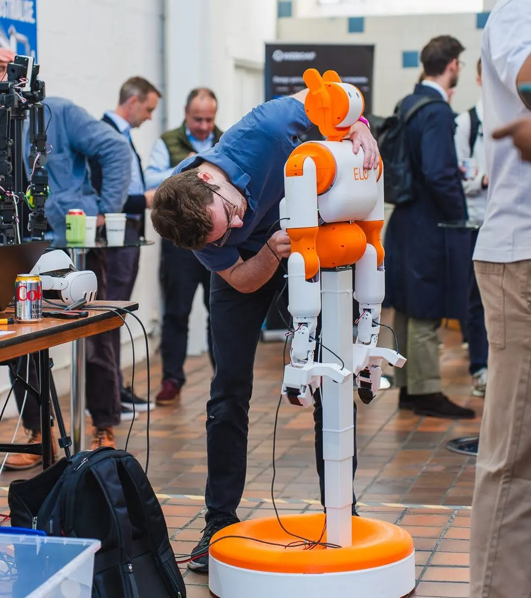 A man in a blue shirt leans over the orange and white Elo humanoid robot on its wheeled stand and adjusts it, with visitors standing and talking on the expo floor behind.