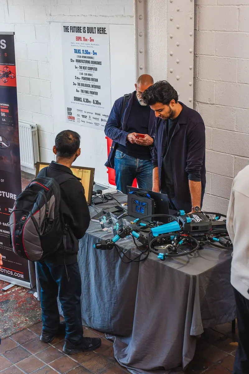 Two men look down at a drone and a laptop on a grey tablecloth at an exhibitor stall, while a third stands in front of the table, under a poster headed "The future is built here" that lists the day's schedule.