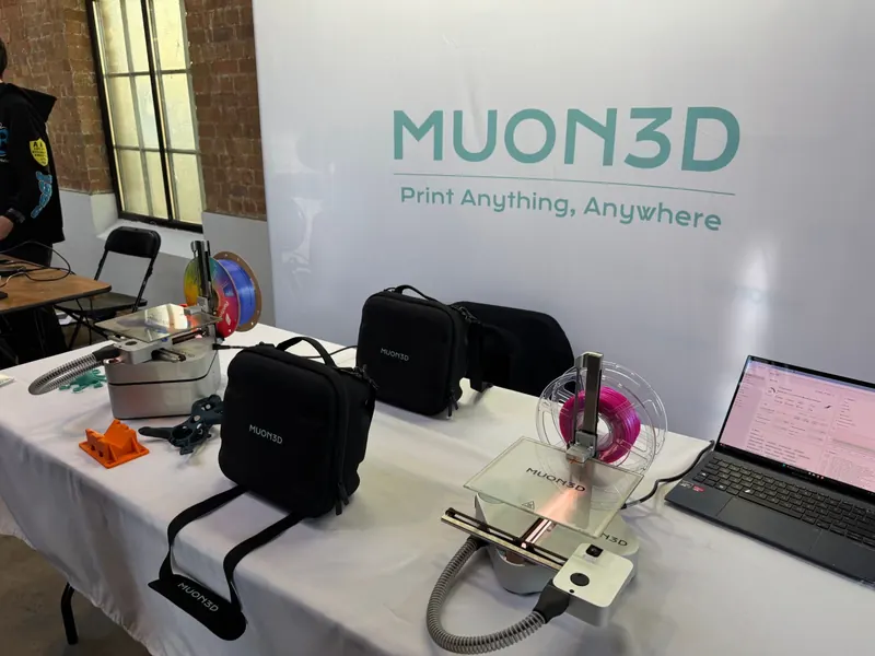 Two small 3D printers and two black carry cases on a white-clothed table, with a laptop on the right, in front of a white Muon3D banner reading "Print Anything, Anywhere".