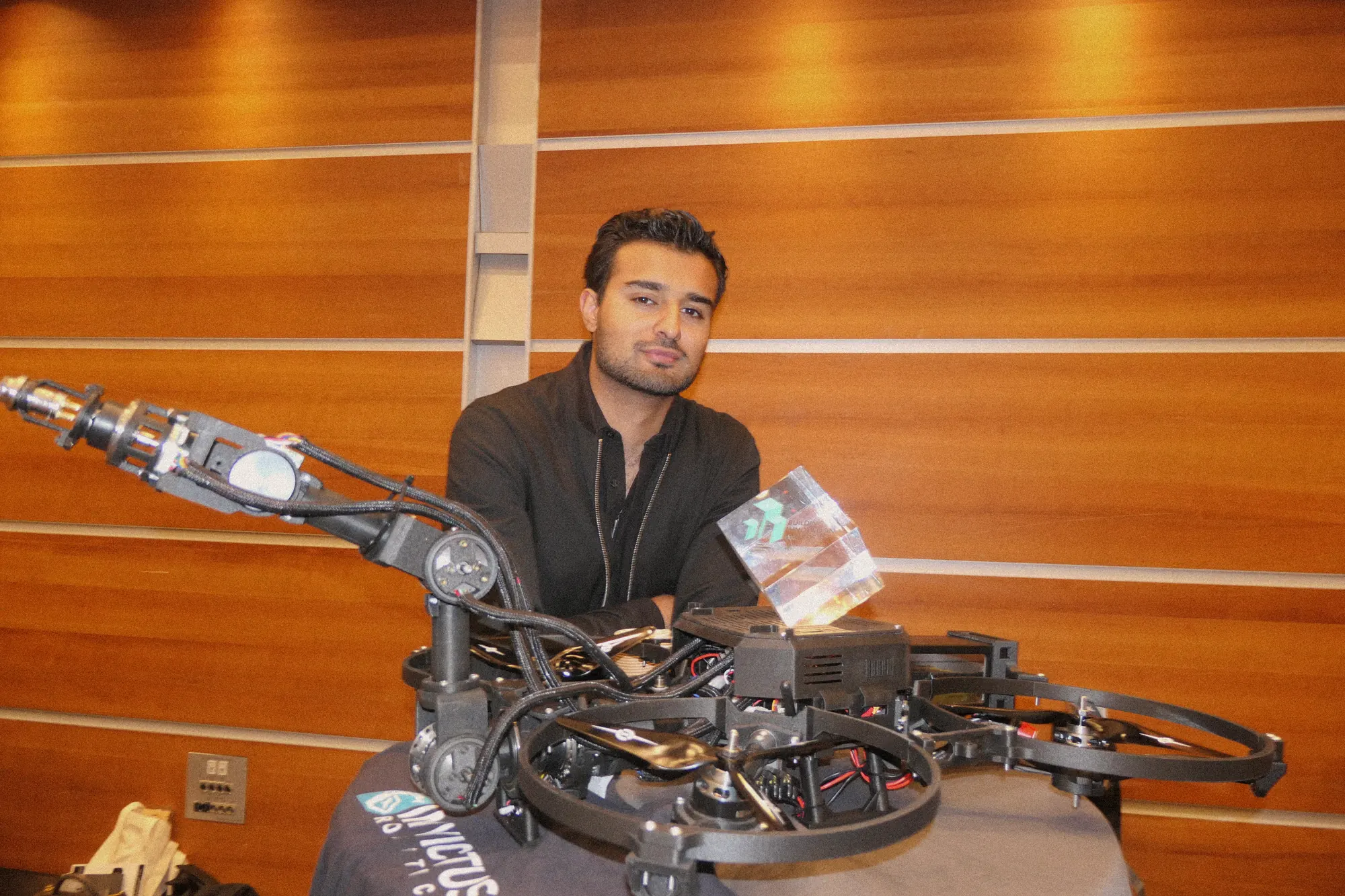 A drone with a robotic arm on a table, its builder behind it.