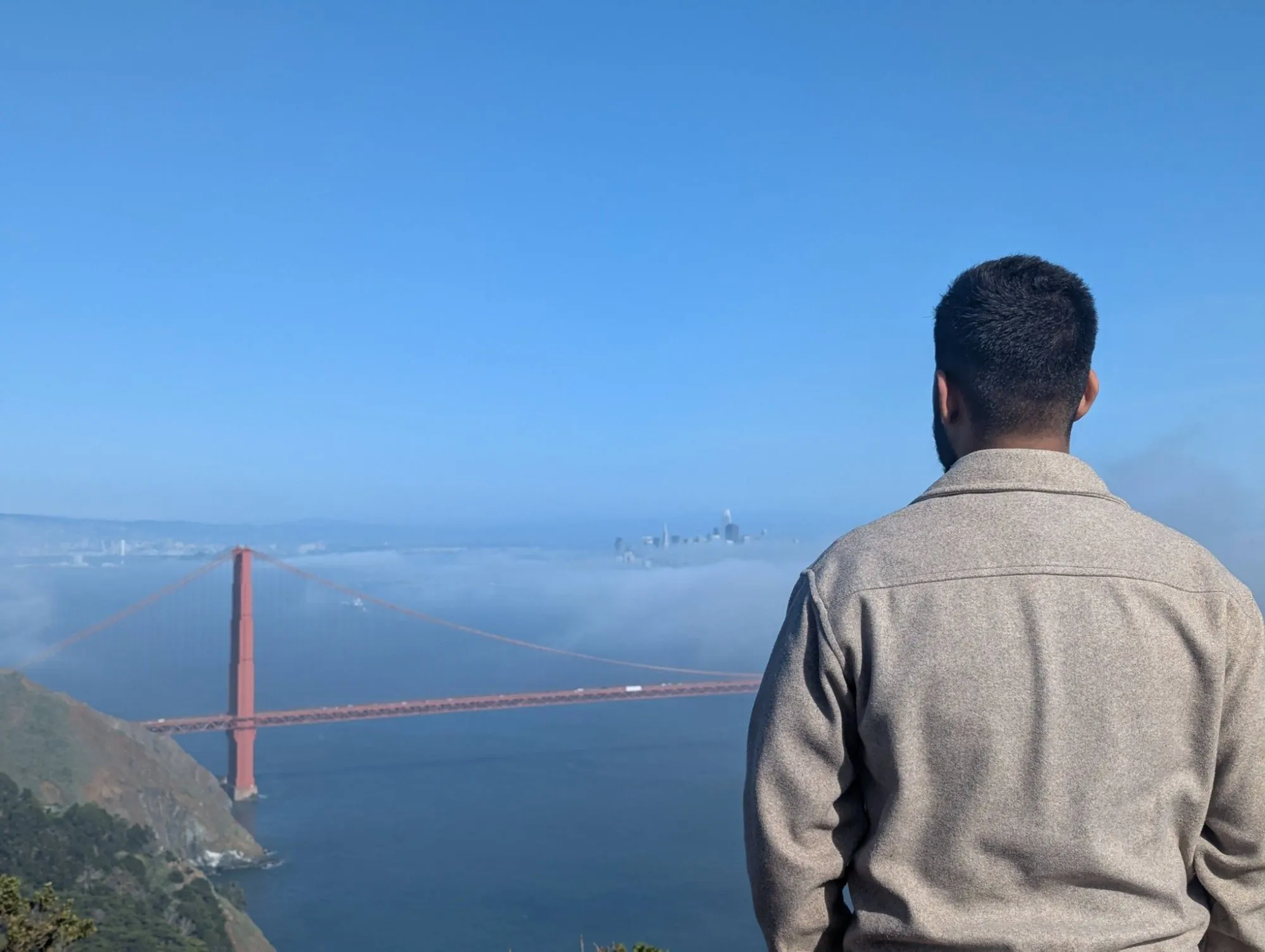 The author, seen from behind in a grey fleece, looks out at the Golden Gate Bridge, its red tower rising out of low fog with the San Francisco skyline faint in the distance.