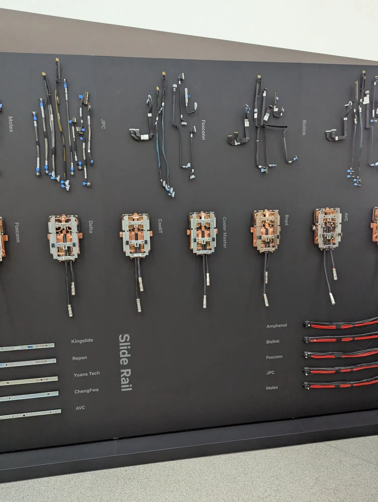 A display wall at Nvidia's headquarters showing server rack components from its suppliers: rows of internal cables, copper liquid-cooling cold plates, slide rails and red power cable harnesses, each labelled with the supplier's name.