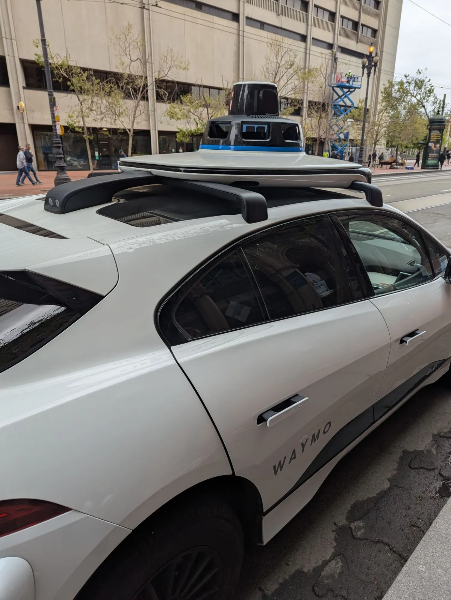A white Waymo robotaxi parked at the kerb, its roof carrying a domed sensor unit, with an office block and trees behind it.