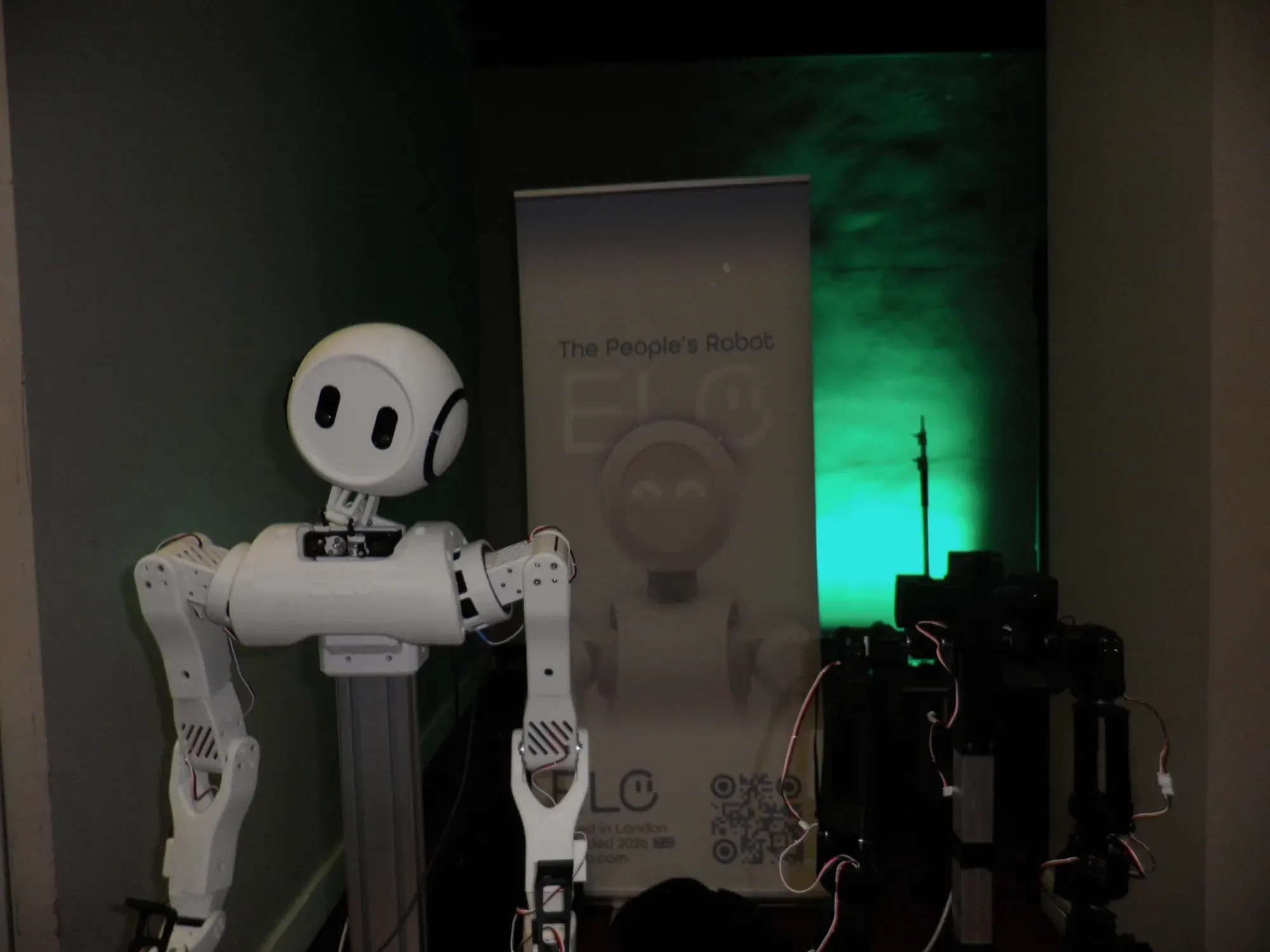 A white wheeled humanoid robot standing in front of the Elo banner, lit green, with other hardware beside it.