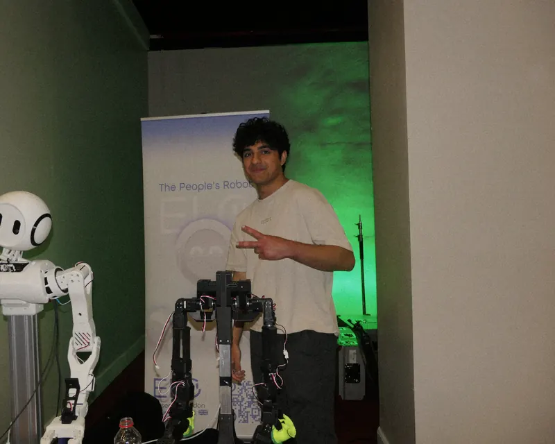 A builder at Muslim Tech Fest making a peace sign beside the Elo humanoid robot and a black robotic arm rig, lit by green stage lights.