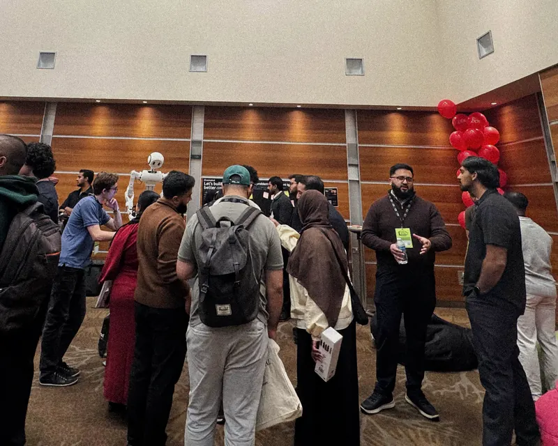 A crowded hall at Muslim Tech Fest, visitors standing in close groups talking, a man with a lanyard in the middle mid-conversation and a humanoid robot behind them.